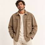A man wearing a brownish-beige button-up shirt jacket over a cream t-shirt and light beige pants, standing against a plain off-white background.