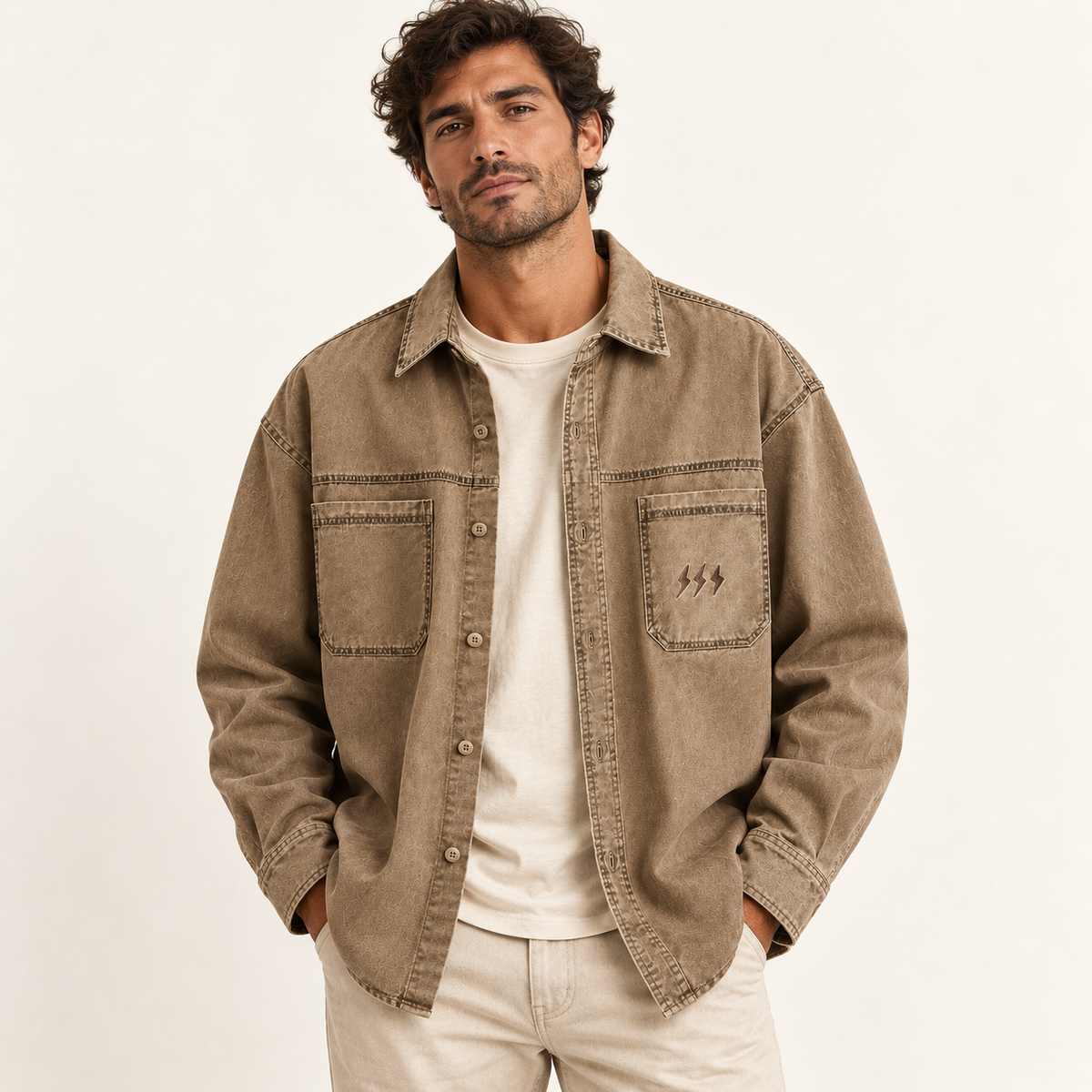 A man wearing a brownish-beige button-up shirt jacket over a cream t-shirt and light beige pants, standing against a plain off-white background.