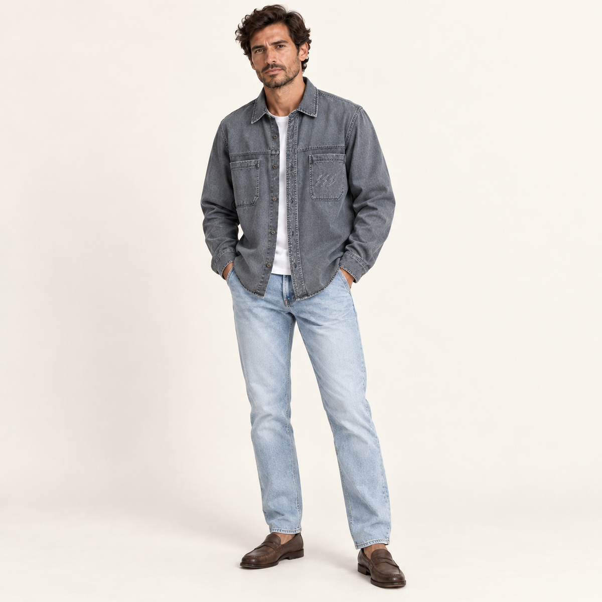 A man modeling a washed gray denim shirt jacket over a white t-shirt, paired with light blue jeans and brown leather loafers, against a plain off-white studio background.